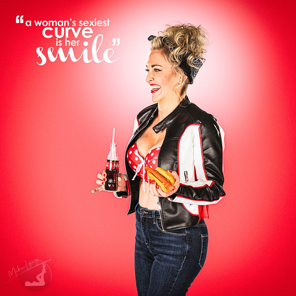 Smiling woman in retro outfit holding a soda and hot dog, standing in front of a pink background with the quote, "a woman's sexiest curve is her smile."